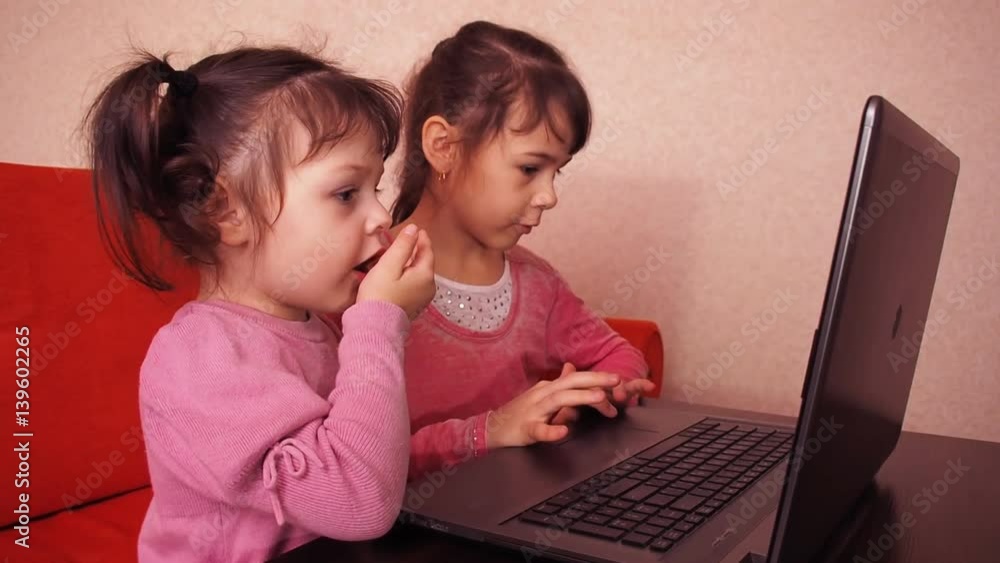 Children playing in the laptop. Two little girls are printed on a laptop. Two sisters sitting on the orange couch looking at a laptop. Close-up. Children playing in the laptop. Two little girls are printed on a laptop. Two sisters sitting on the orange couch looking at a laptop. Close-up.