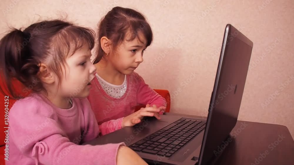 Children playing in the laptop. Two little girls are printed on a laptop. Two sisters sitting on the orange couch looking at a laptop. Close-up. Children playing in the laptop. Two little girls are printed on a laptop. Two sisters sitting on the orange couch looking at a laptop. Close-up.