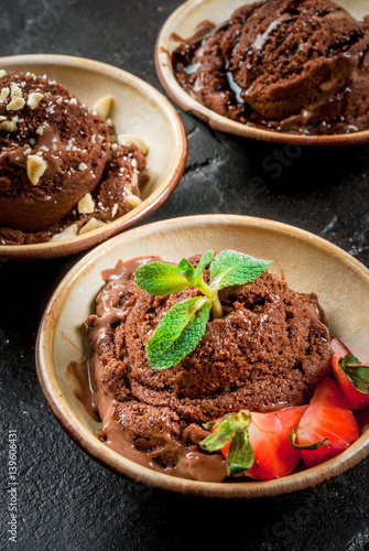 Three kinds of chocolate ice cream decoration: with chocolate sauce (syrup), with slices of strawberry and mint, with pieces of chopped nuts. On the black concrete stone table