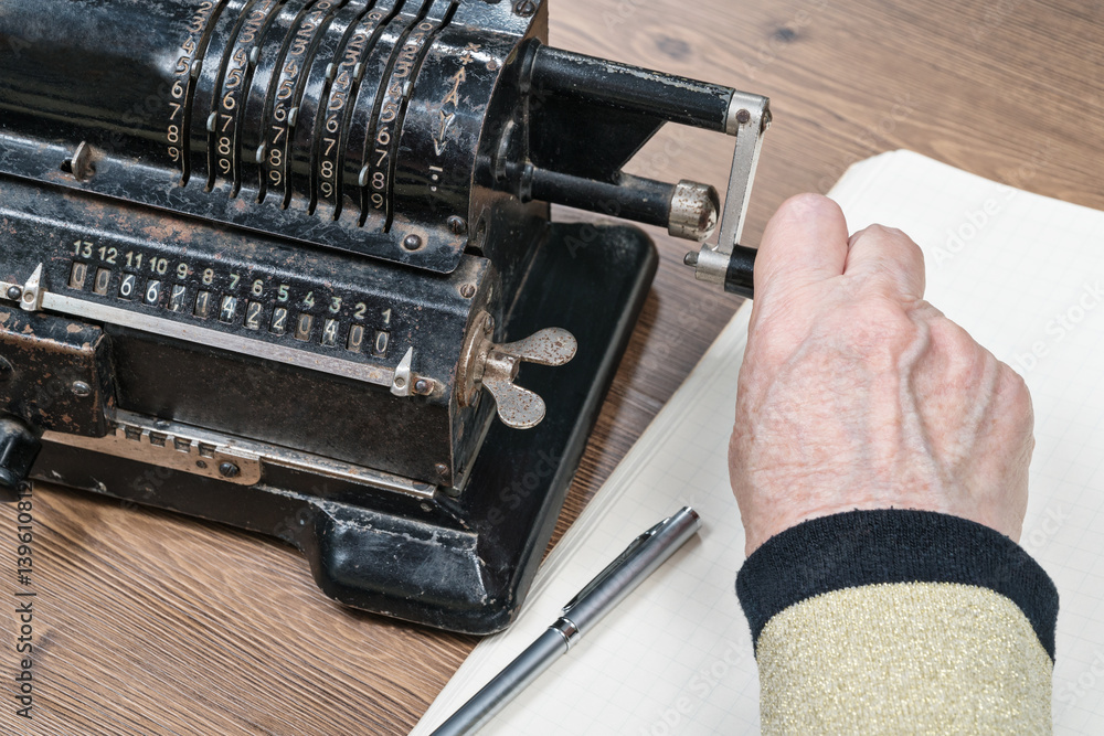 Old wrinkled hand turns the handle retro adding machine lying on the table next to a sheet of paper and a pen, close-up Old wrinkled hand turns the handle retro adding machine lying on the table next to a sheet of paper and a pen, close-up