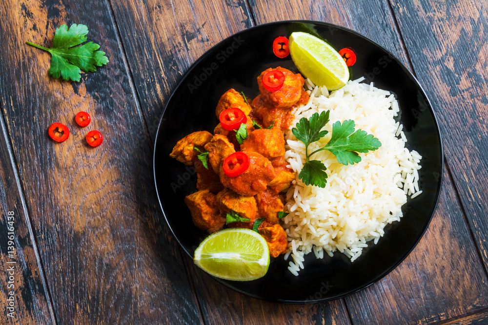 Traditional Indian cuisine, chicken curry with fresh chilli pepper, lime and parsley and boiled rice on a black ceramic plate on the wooden table, top view.