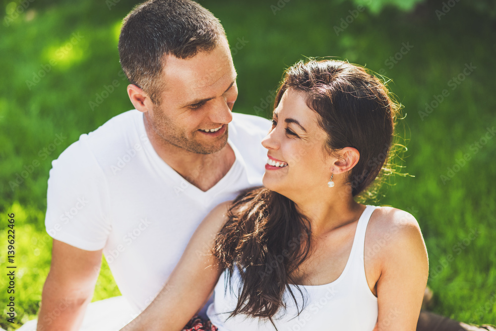 Close up portrait of beautiful couple in love sitting on a picnic blanket outdoor. Caucasian happy dreamy man and smiling brunette woman sitting on green grass in park looking to each other.. Close up portrait of beautiful couple in love sitting on a picnic blanket outdoor. Caucasian happy dreamy man and smiling brunette woman sitting on green grass in park looking to each other..