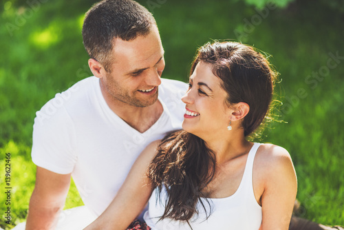 Close up portrait of beautiful couple in love sitting on a picnic blanket outdoor. Caucasian happy dreamy man and smiling brunette woman sitting on green grass in park looking to each other..