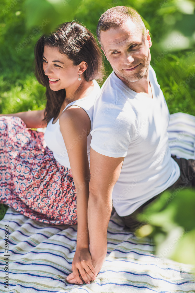 Fototapeta premium Vertical top view of a attractive couple in love sitting on a picnic blanket. Caucasian happy dreamy man and smiling brunette woman looking away on plaid on green grass.