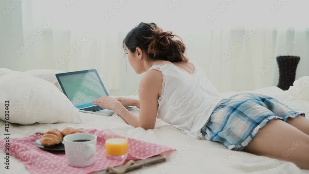 Young woman using laptop during breakfast lying on white bed at home. Brunette girl typing on pc computer. Young woman using laptop during breakfast lying on white bed at home. Brunette girl typing on pc computer.