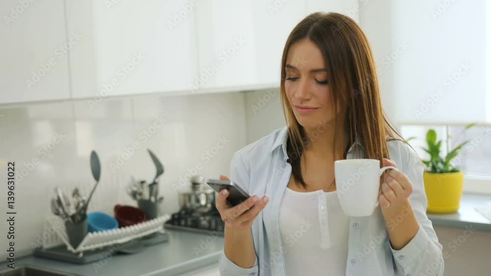 Pretty woman using smart phone, texting and drinking coffee in the kitchen. Pretty woman using smart phone, texting and drinking coffee in the kitchen.