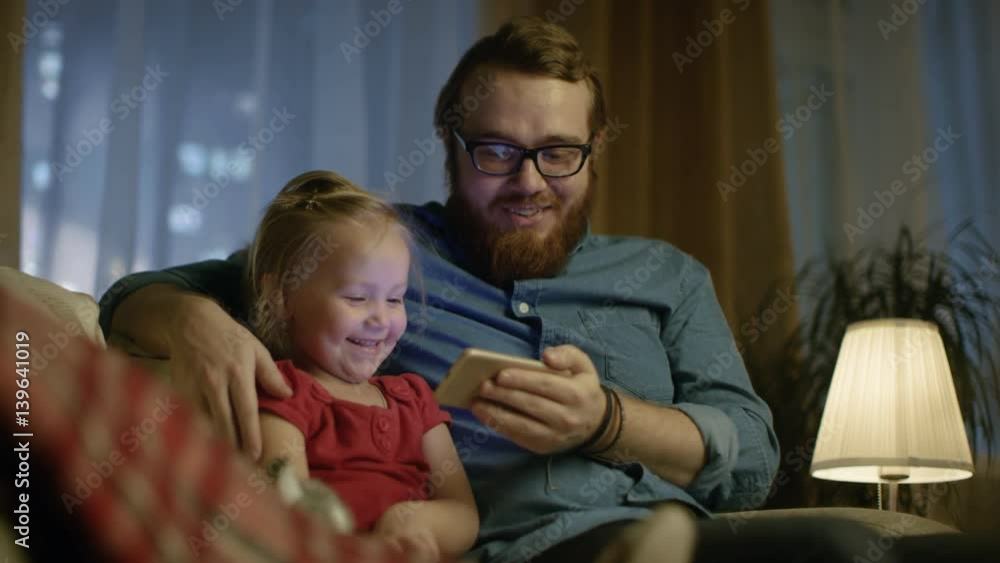 In the Evening Father and Little Daughter Sitting on a Sofa in the Living Room Watch Funny Videos on Smartphone. Shot on RED EPIC-W 8K Helium Cinema Camera. In the Evening Father and Little Daughter Sitting on a Sofa in the Living Room Watch Funny Videos on Smartphone. Shot on RED EPIC-W 8K Helium Cinema Camera.