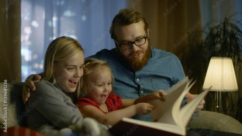Father, Mother and Little Daughter Reading Children's Book on a Sofa in the Living Room. It's Evening. Shot on RED EPIC-W 8K Helium Cinema Camera.