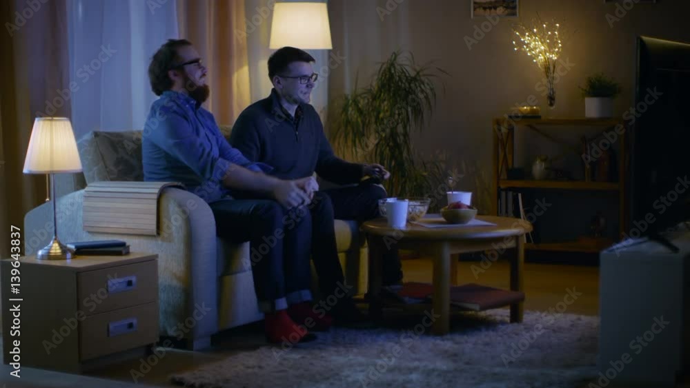 In the Evening Two Friends are Sitting on a Sofa in the Living Room and Playing Competitive Video Games. One of Them Wins and He is Enjoying His Success. Shot on RED EPIC-W 8K Helium Cinema Camera. In the Evening Two Friends are Sitting on a Sofa in the Living Room and Playing Competitive Video Games. One of Them Wins and He is Enjoying His Success. Shot on RED EPIC-W 8K Helium Cinema Camera.