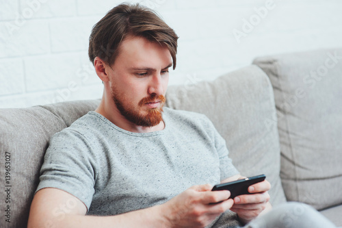 The bearded man sitting on the couch at home and playing the phone