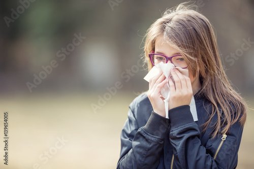 Girl with allergy symptom blowing nose. Teen girl using a tissue in a park.