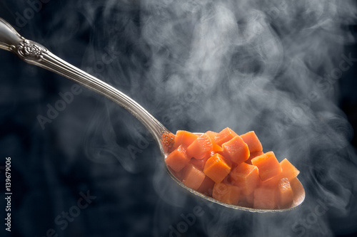 Carrot cubes in a spoon with water vapor. Vegetarian cuisine. Dark background. Cooking food.