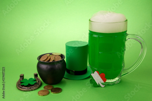 St Patrick's Day green beer with shamrock, pot with gold coins, horseshoe and Leprechaun hat against green background.