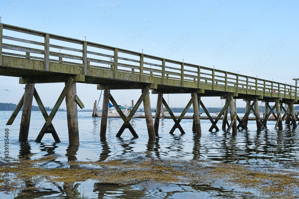 Close view of a very long pier over water with seaweed in the foreground and sailboats in the distance at Stockton Springs Maine in the summertime.