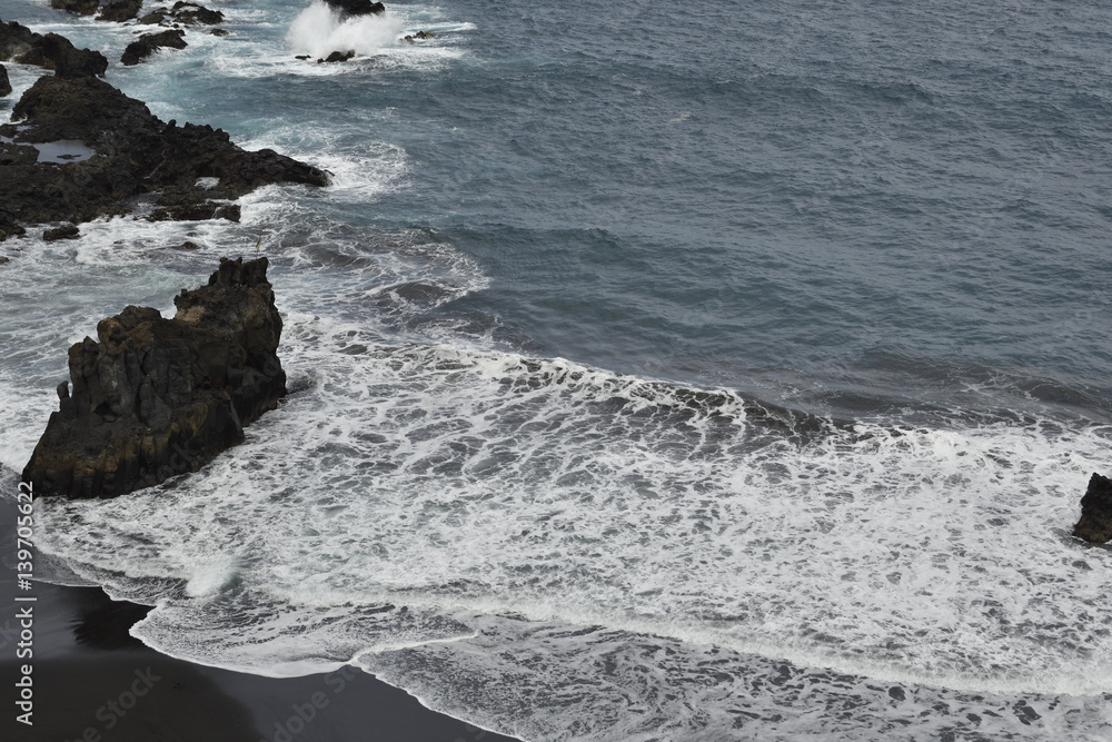 Waves against a beach with black lava sand and cliffs