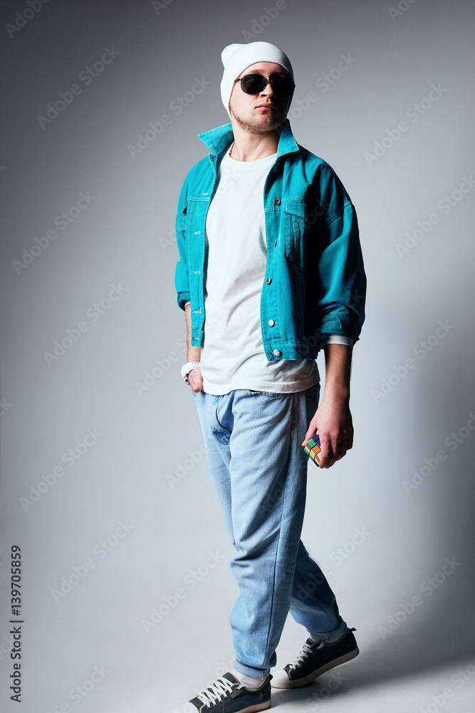 Male fashion concept. Fashionable young man wearing trendy posing over gray background. Hands in pockets, crossed legs. Street style. Studio shot