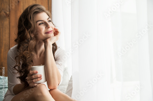 Cheerful young lady holding white mug and looking at window. Horizontal indoors shot.