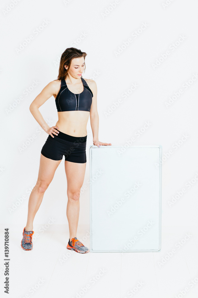 Happy smiling sports woman in black sportswear showing blank signboard, over white wall Happy smiling sports woman in black sportswear showing blank signboard, over white wall