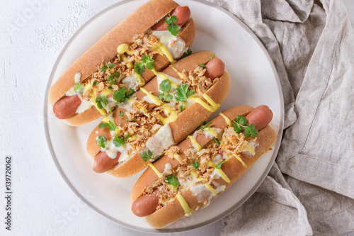 Hot dogs with sausage, fried onion, coriander leaves, cheese sauce and mustard, served on white ceramic plate with textile over white concrete texture background. Fast food. Top view, copy space