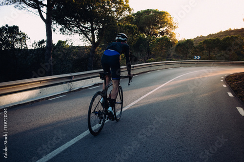 A fit young cyclist riding into the sunset on an empty dark road © onewithahalf