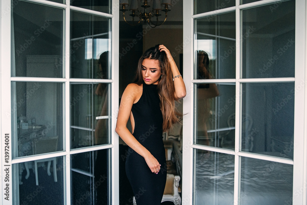 Attractive charming sexy girl in stylish black dress in hotel room near the door of balcony Attractive charming sexy girl in stylish black dress in hotel room near the door of balcony