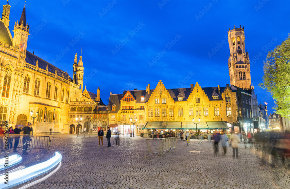 Naklejka premium BRUGES, BELGIUM - MARCH 2015: Tourists visit ancient medieval Burg Square at night. Brugge attracts more than 2 million people annually