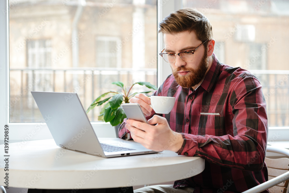 Bearded young man drinking coffee while using laptop and phone. Bearded young man drinking coffee while using laptop and phone.