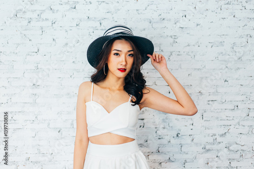 Young happy Asian woman smiling in fashionable dress and summer hat over white brick wall