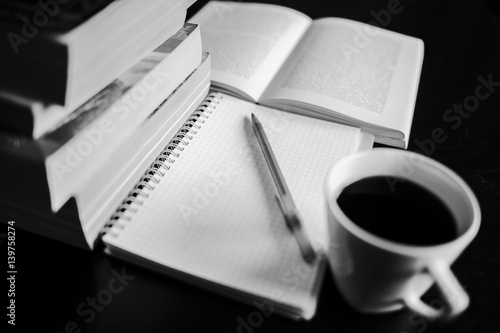 There are coffee, books, notebook and pen. Note-taking. Black and white photo.