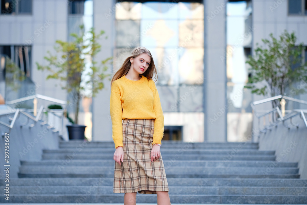 Blond hair young woman in yellow jamper and check skirt posing and against modern building. Street fashion style concept. 