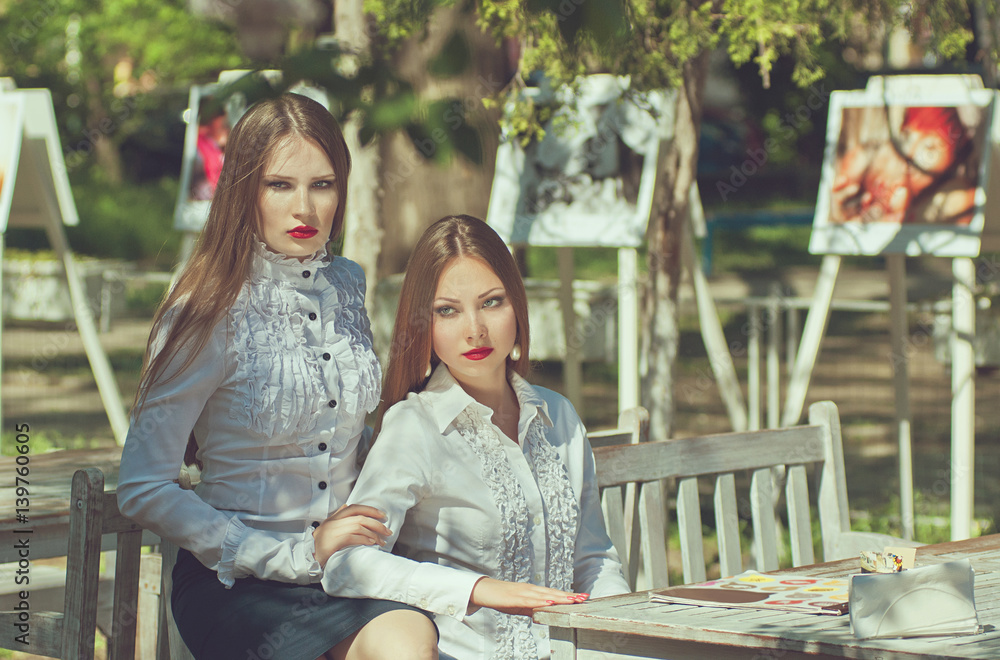 Beautiful portrait of two serious young woman, elegantly dressed in white shirts and dark skirts, sitting in park on wooden bench at a table in a warm sunny day Beautiful portrait of two serious young woman, elegantly dressed in white shirts and dark skirts, sitting in park on wooden bench at a table in a warm sunny day