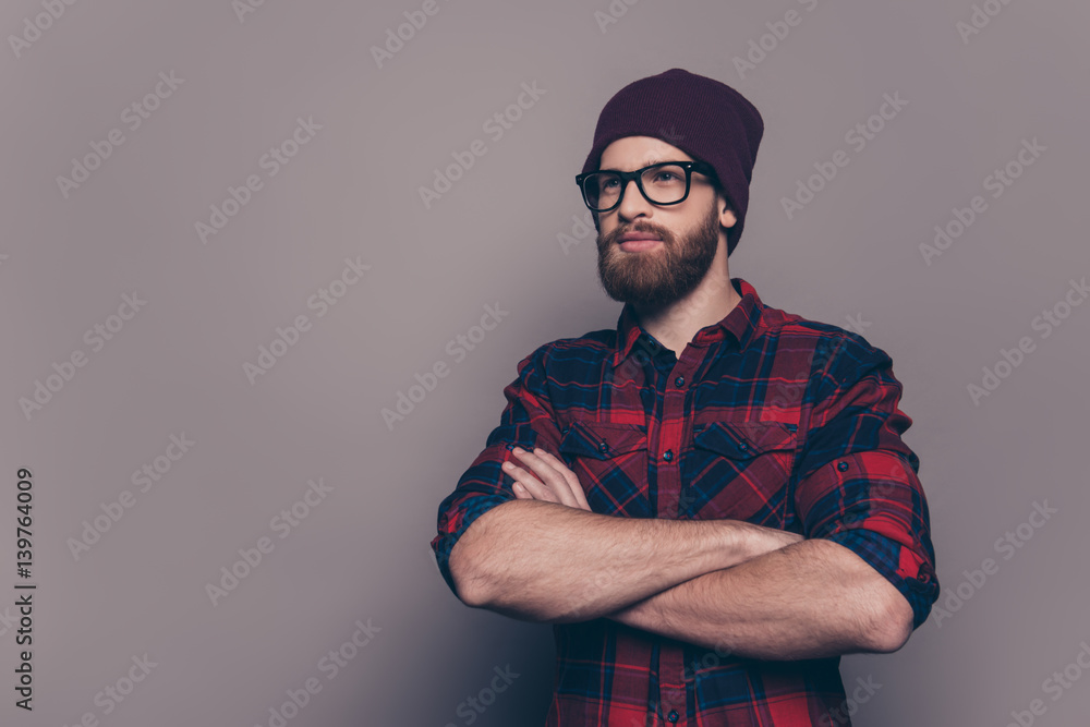 A portrait of young stylish guy wearing checkered shirt, glasses and hat standing with crossed hands in front of gray background A portrait of young stylish guy wearing checkered shirt, glasses and hat standing with crossed hands in front of gray background