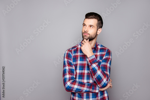 A portrait of young lost in thought freelancer touching his chin and looking up against gray background