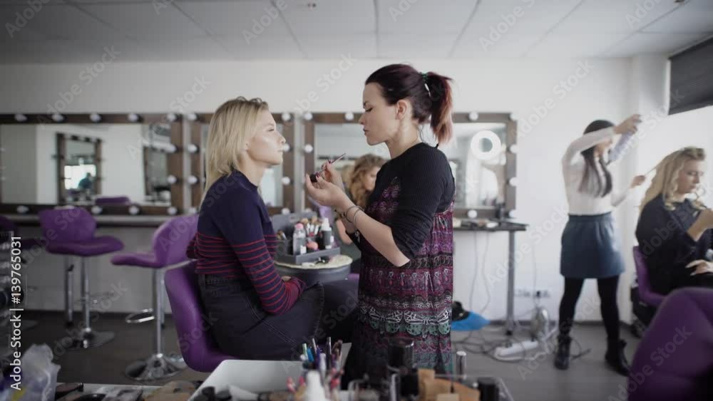 Make-up artist is working preparing model for future shooting. Brunette female master is applying cosmetics on client's face while hairdresser is finishing hairdo for blonde woman in the background. Make-up artist is working preparing model for future shooting. Brunette female master is applying cosmetics on client's face while hairdresser is finishing hairdo for blonde woman in the background.
