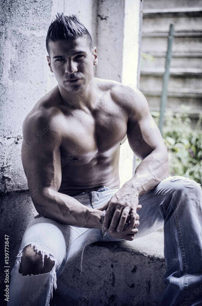 Muscular young latino man shirtless in jeans sitting against concrete wall looking at camera