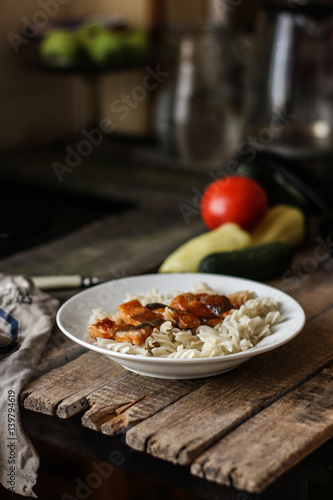 Fish in tomato sauce and pasta in a white plate on a wooden surface (on a table) and vegetables