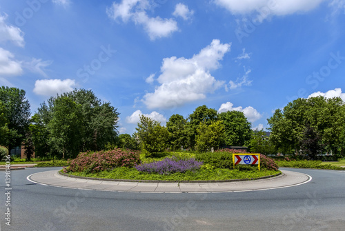 Small roundabout with plants, trees and traffic sign without any traffic, against a beautiful blue sky