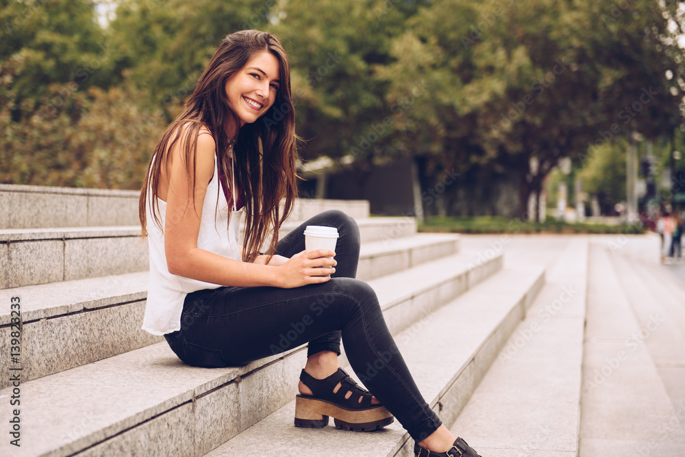 Smiling young woman with the paper coffee cup sitting on stairs and looking at camera. Smiling young woman with the paper coffee cup sitting on stairs and looking at camera.