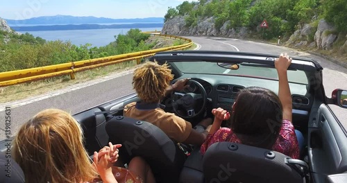 Friends driving on the motorway in red convertible and pointing out the sea