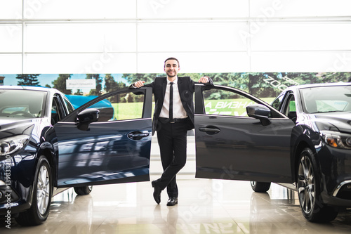 Confident smiling car salesman at the showroom near two cars, he is standing near open cars doors of luxury cars