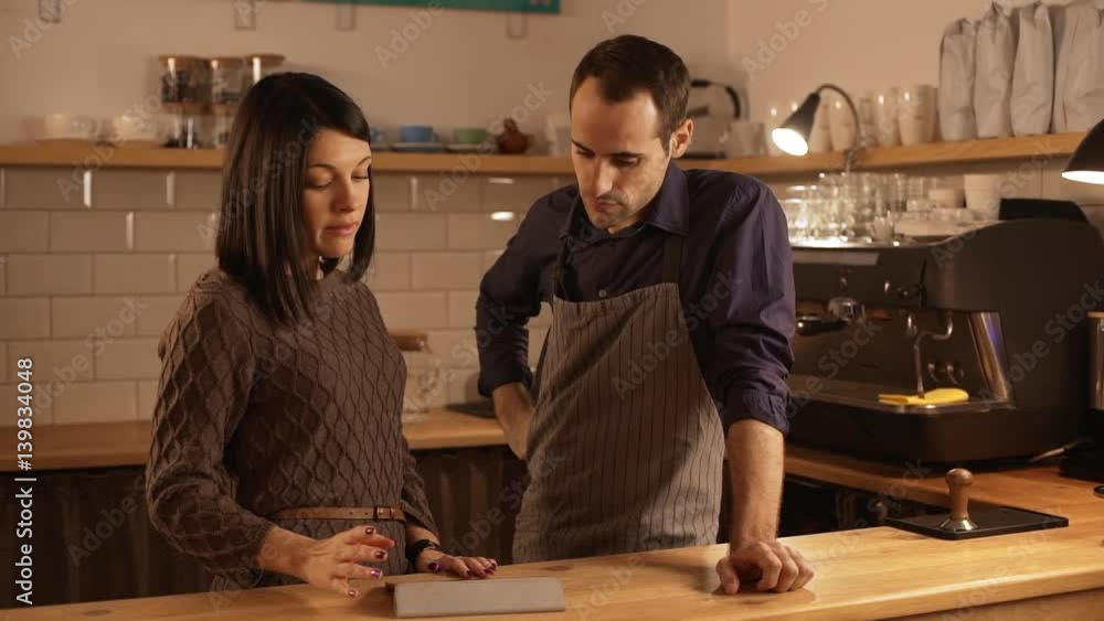 Caucasian brunette female and male middle-aged barista wearing apron standing behind the counter and talking in slowmotion Caucasian brunette female and male middle-aged barista wearing apron standing behind the counter and talking in slowmotion
