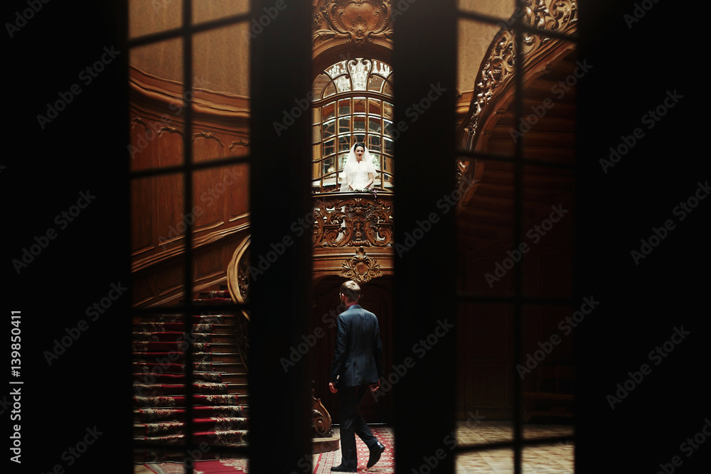 elegant gorgeous bride and stylish groom standing on wooden stairs in rich room. unusual view through doors. luxury wedding concept in retro style