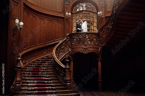 elegant gorgeous bride and stylish groom standing on wooden stairs in amazing old rich room. unusual wedding couple in retro style. luxury wedding concept