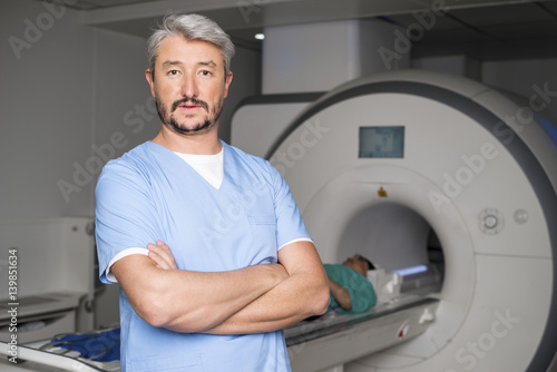 Doctor Standing Arms Crossed With Patient On CT Scan Machine