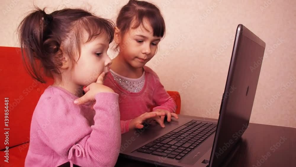 Children playing in the laptop. Two little girls are printed on a laptop. Two sisters sitting on the orange couch looking at a laptop. Close-up. Children playing in the laptop. Two little girls are printed on a laptop. Two sisters sitting on the orange couch looking at a laptop. Close-up.