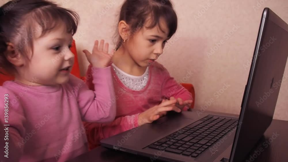 Children playing in the laptop. Two little girls are printed on a laptop. Two sisters sitting on the orange couch looking at a laptop. Close-up. Children playing in the laptop. Two little girls are printed on a laptop. Two sisters sitting on the orange couch looking at a laptop. Close-up.