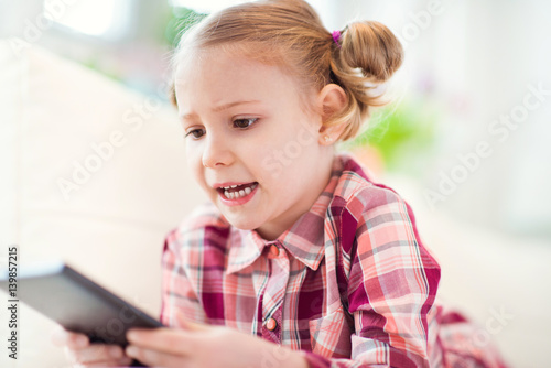 Pretty little child girl using a digital tablet, looking and smiling while lying on the whitw couch