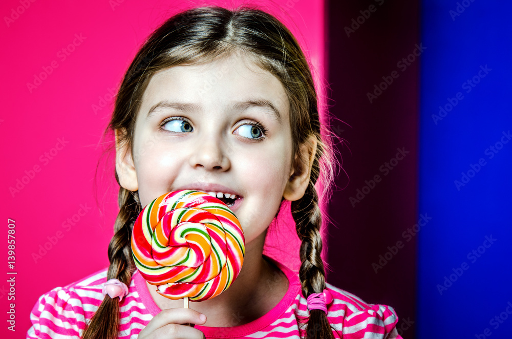 Portrait of a little girl with beautiful big eyes. The girl is holding a large multi-colored candy and licks it.