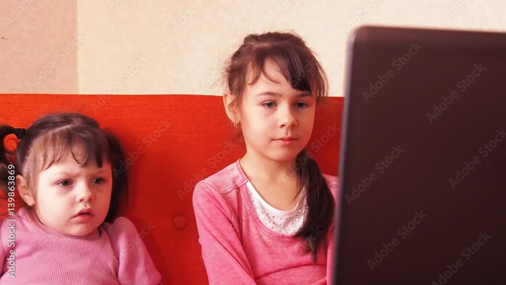 Children playing in the laptop. Two little girls are printed on a laptop. Two sisters sitting on the orange couch looking at a laptop. Close-up. Children playing in the laptop. Two little girls are printed on a laptop. Two sisters sitting on the orange couch looking at a laptop. Close-up.