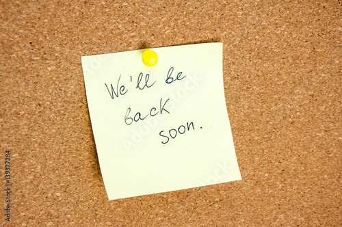 Paper note written with WE'LL BE BACK SOON  inscription on cork board